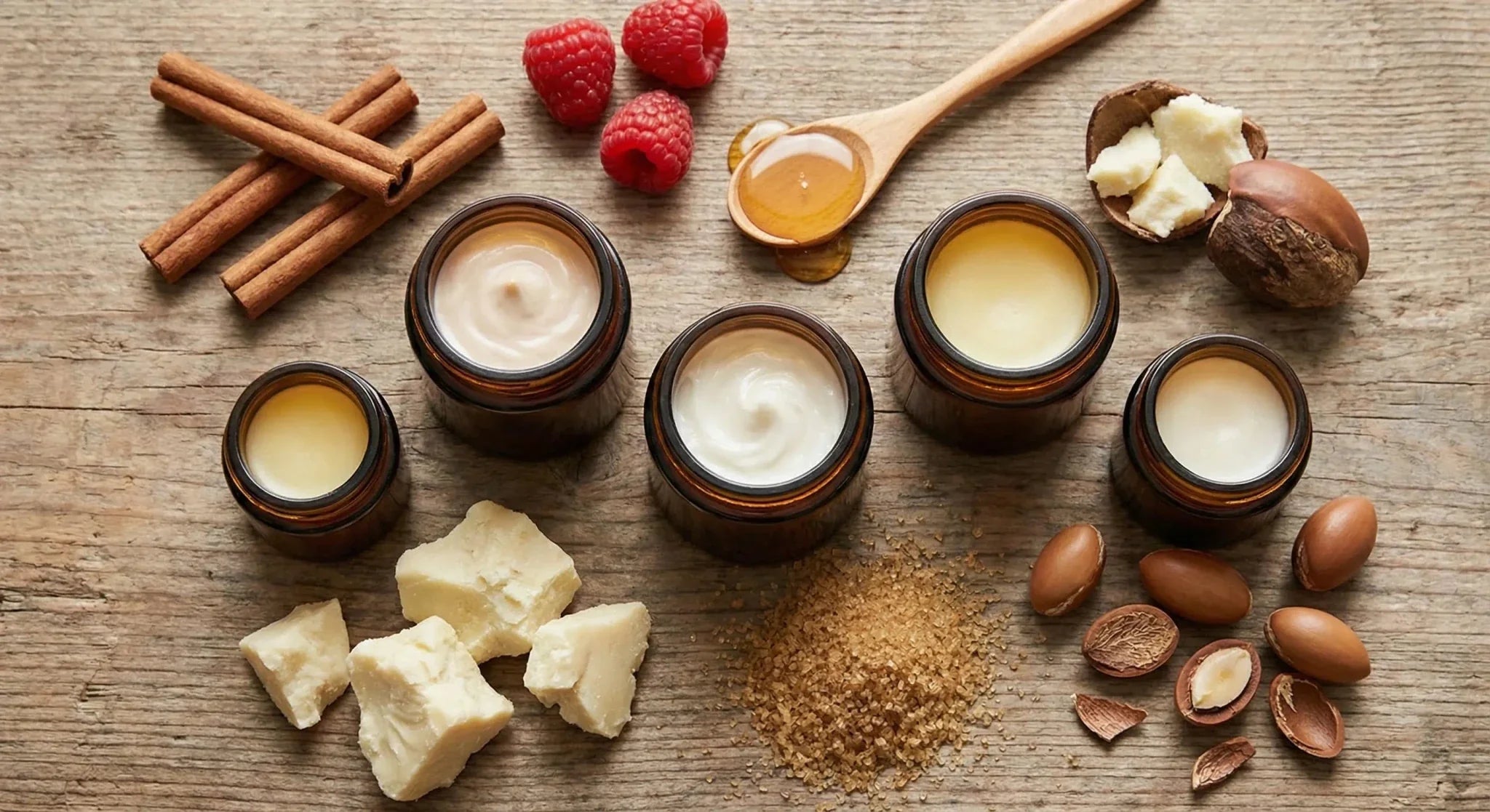 Cinq pots de baume à lèvres entourés de leurs ingrédients : beurre de karité, miel, cannelle, framboise, sucre et noix d'argan