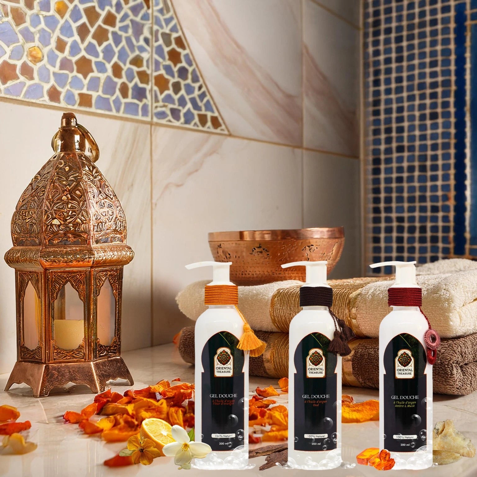 Dans un hammam au style marocain on voit trois gel douche de la marque oriental treasure posé sur un plan en marbre clair avec de l'ambre, du musk, du oud et des fleurs d'oranger