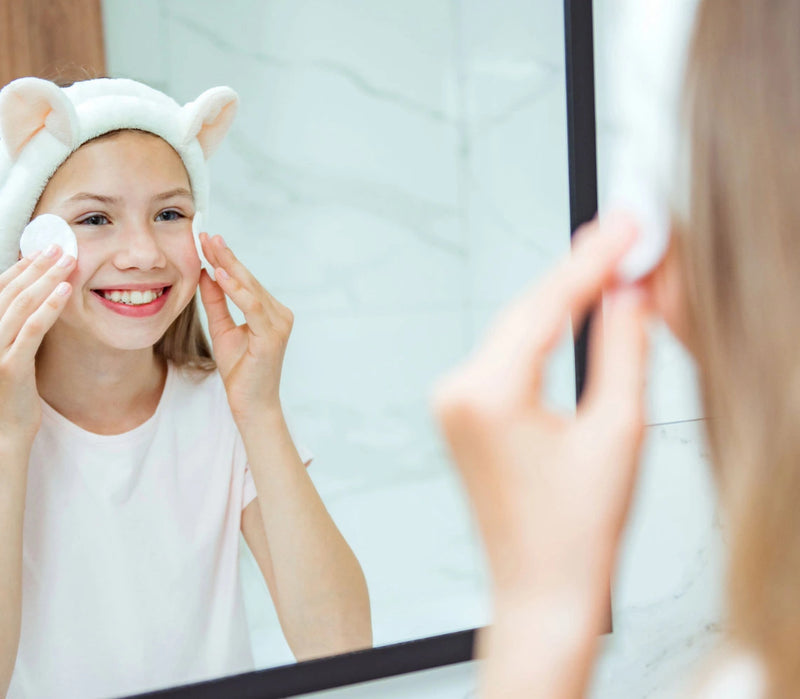 Une jeune fille souriante devant un miroir applique ses produits pour sa routine peau adolescente